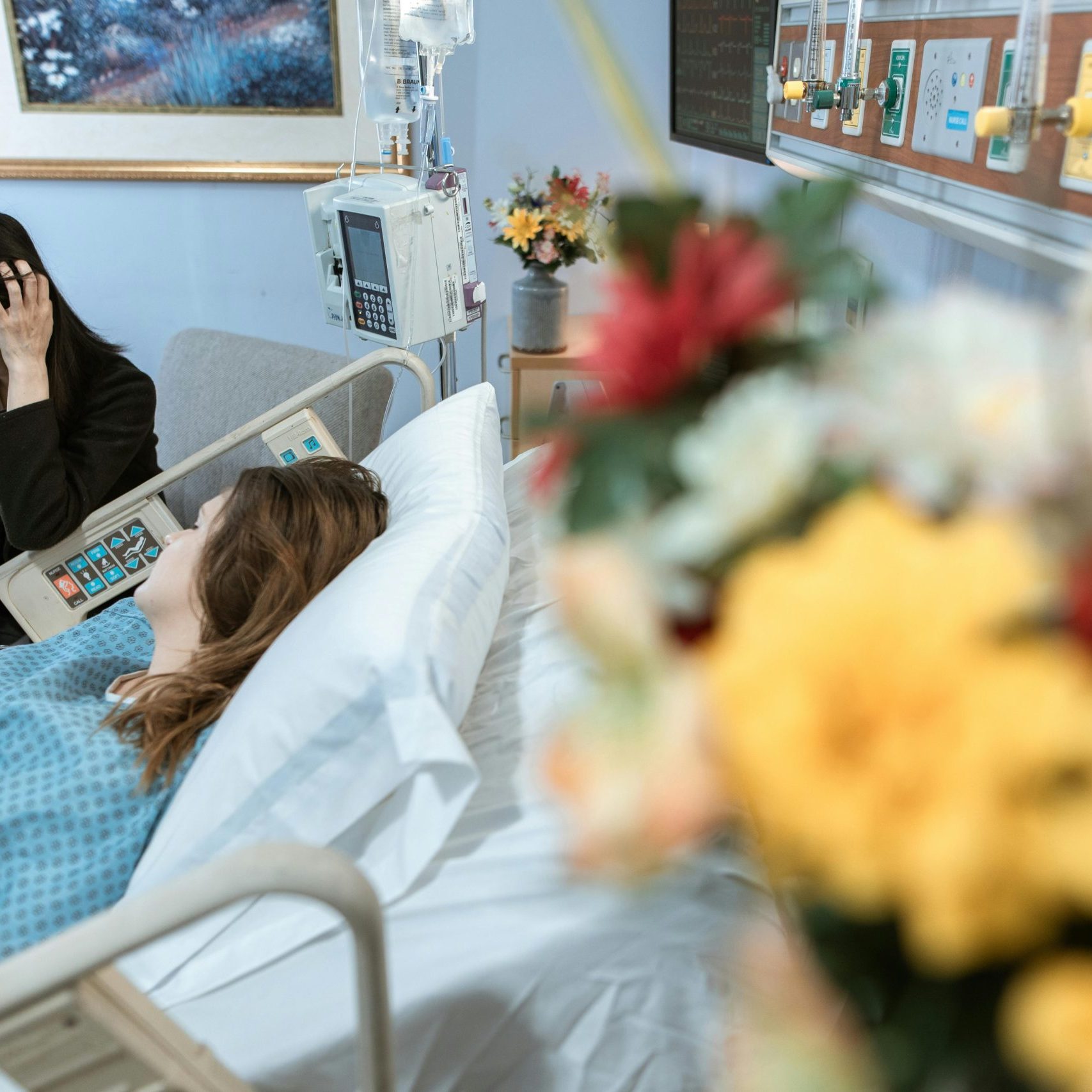 Eine Frau liegt in einem Krankenhausbett und wird besucht, Blumen im Vordergrund – © RDNE Stock project/Pexels